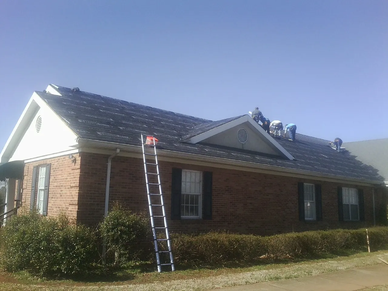 Crew tearing shingles off a brick ranch roof with a ladder set — Metal Roof Repair work by our crew in Tonica