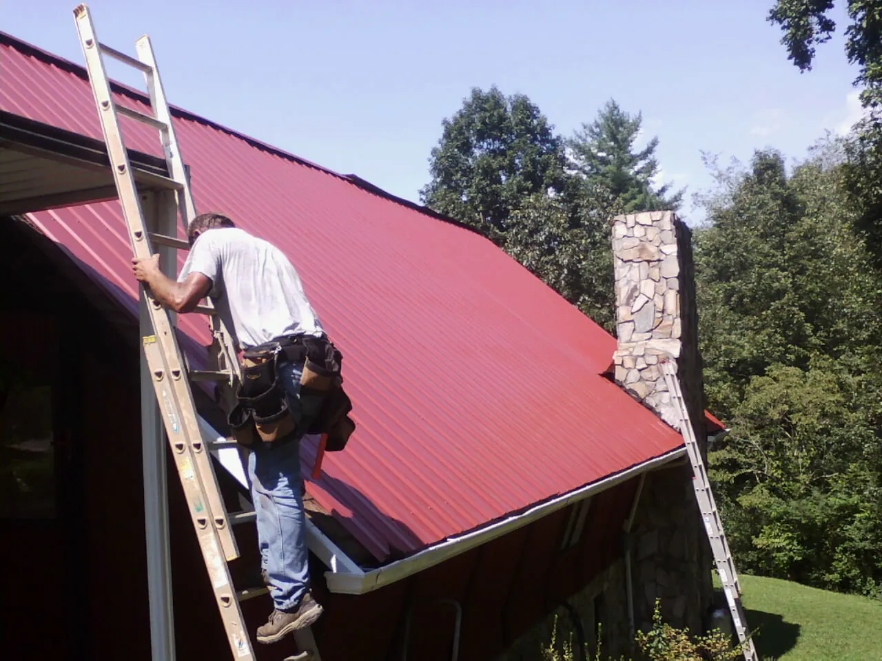 Roofer on a ladder beside a red metal roof and stone chimney — Soffit & Fascia Repair by Roofing Brothers in Tonica
